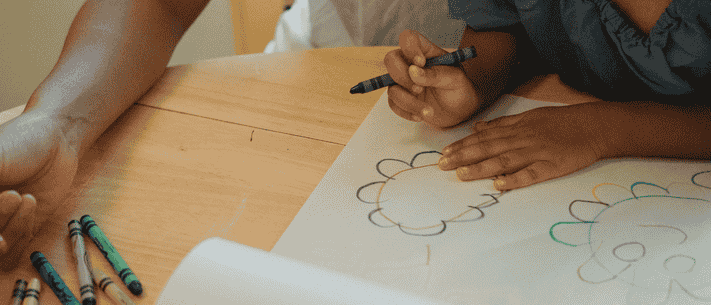 Adult And Child Drawing A Flower Together With Crayons On Paper At A Wooden Table