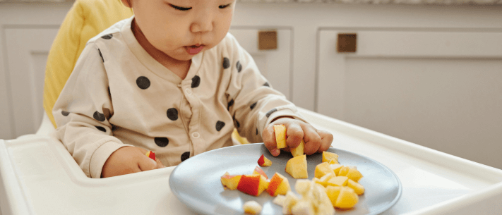 Baby in High Chair Holding Fruit