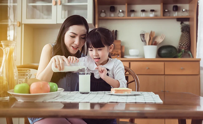 Mom guiding preschool daughter as she pours milk into a glass in the dining area
