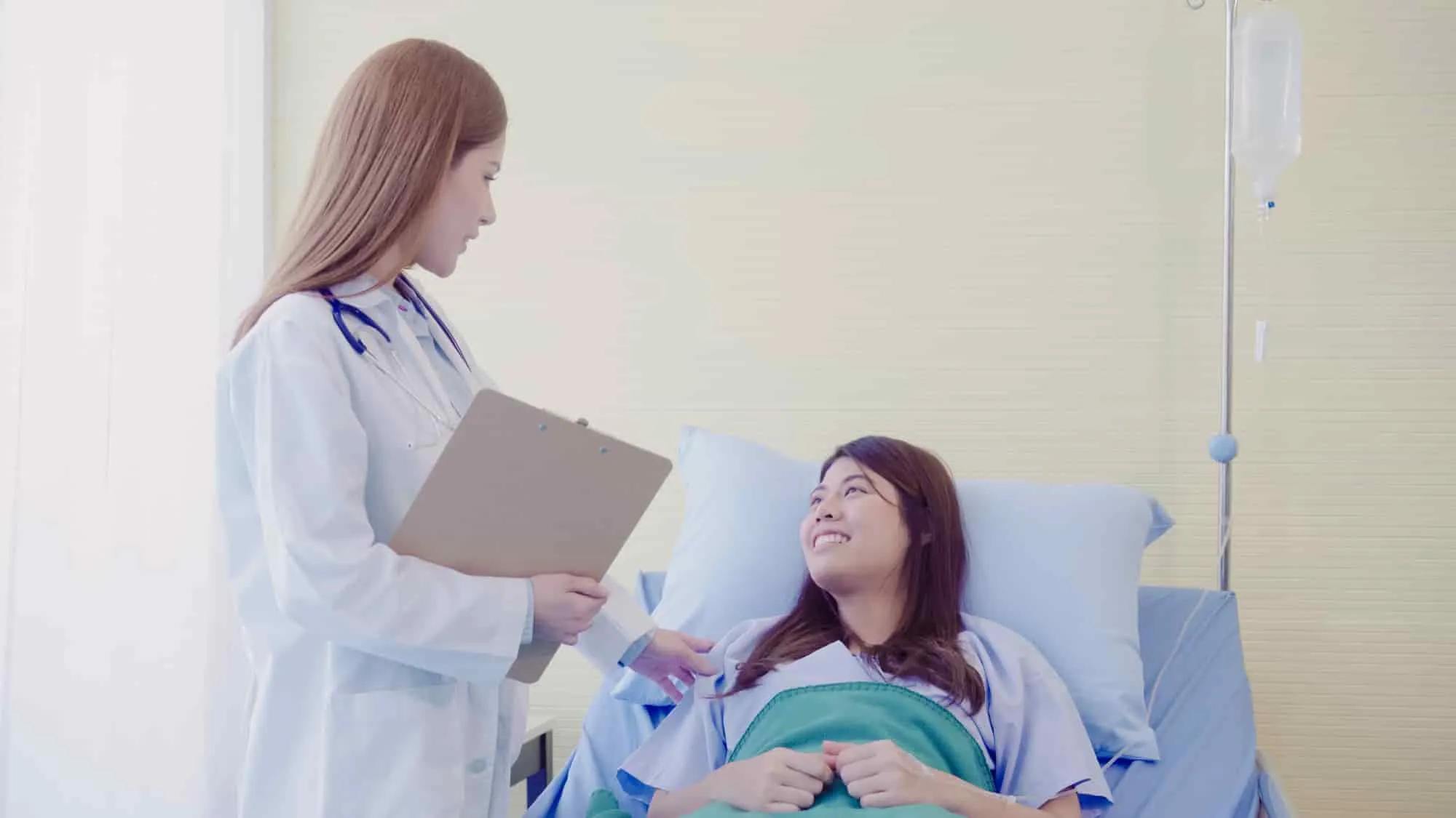 Doctor with a clipboard consulting a smiling mother after labor in a hospital bed.