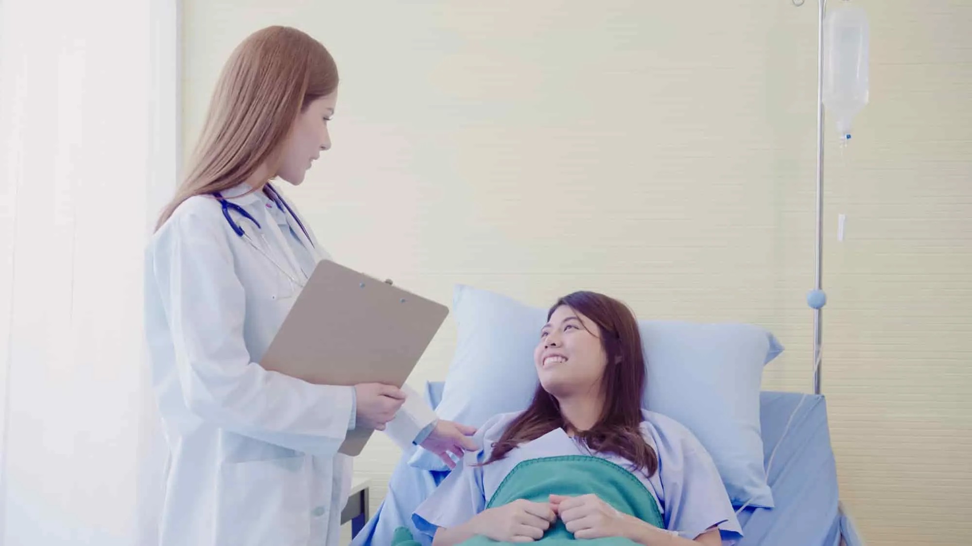 Doctor with a clipboard consulting a smiling mother after labor in a hospital bed.