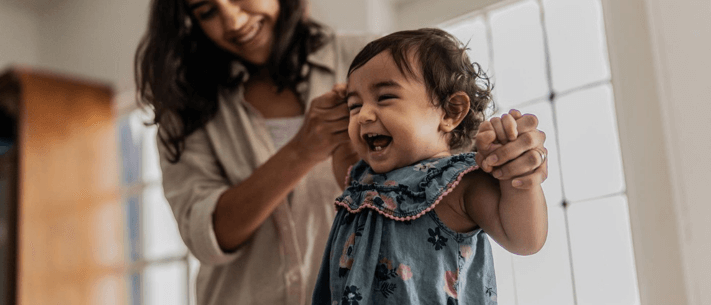 Smiling Baby Girl in Floral Denim Dress with Mother Behind Indoors
