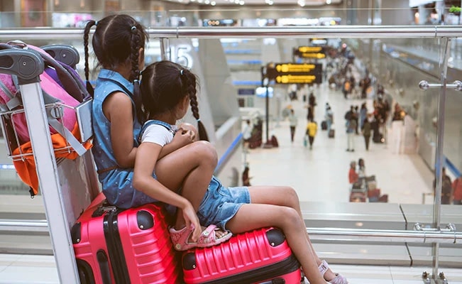 Two Young Girls With Pigtails Sitting On Pink Luggage At Airport Wearing Denim Dresses