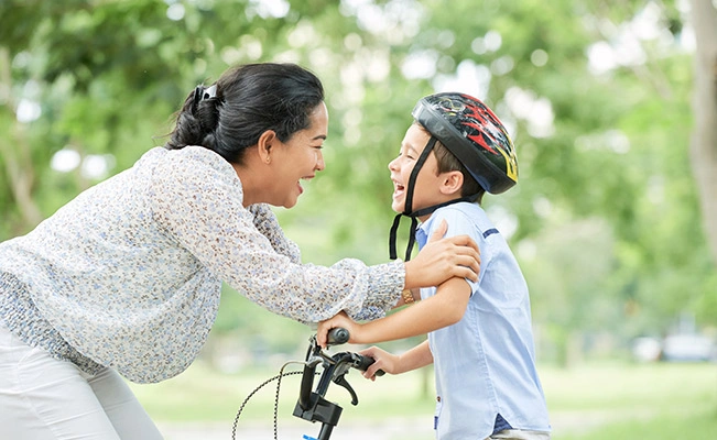 Mother Smiling At Her Son On A Bicycle While Holding His Arms, Outdoors With Blurry Green And White Background