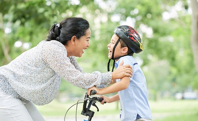 Mother Smiling At Her Son On A Bicycle While Holding His Arms, Outdoors With Blurry Green And White Background