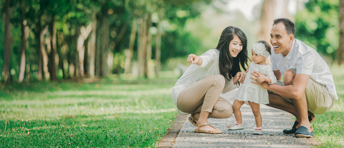 Parents Helping Toddler Take First Steps In Park