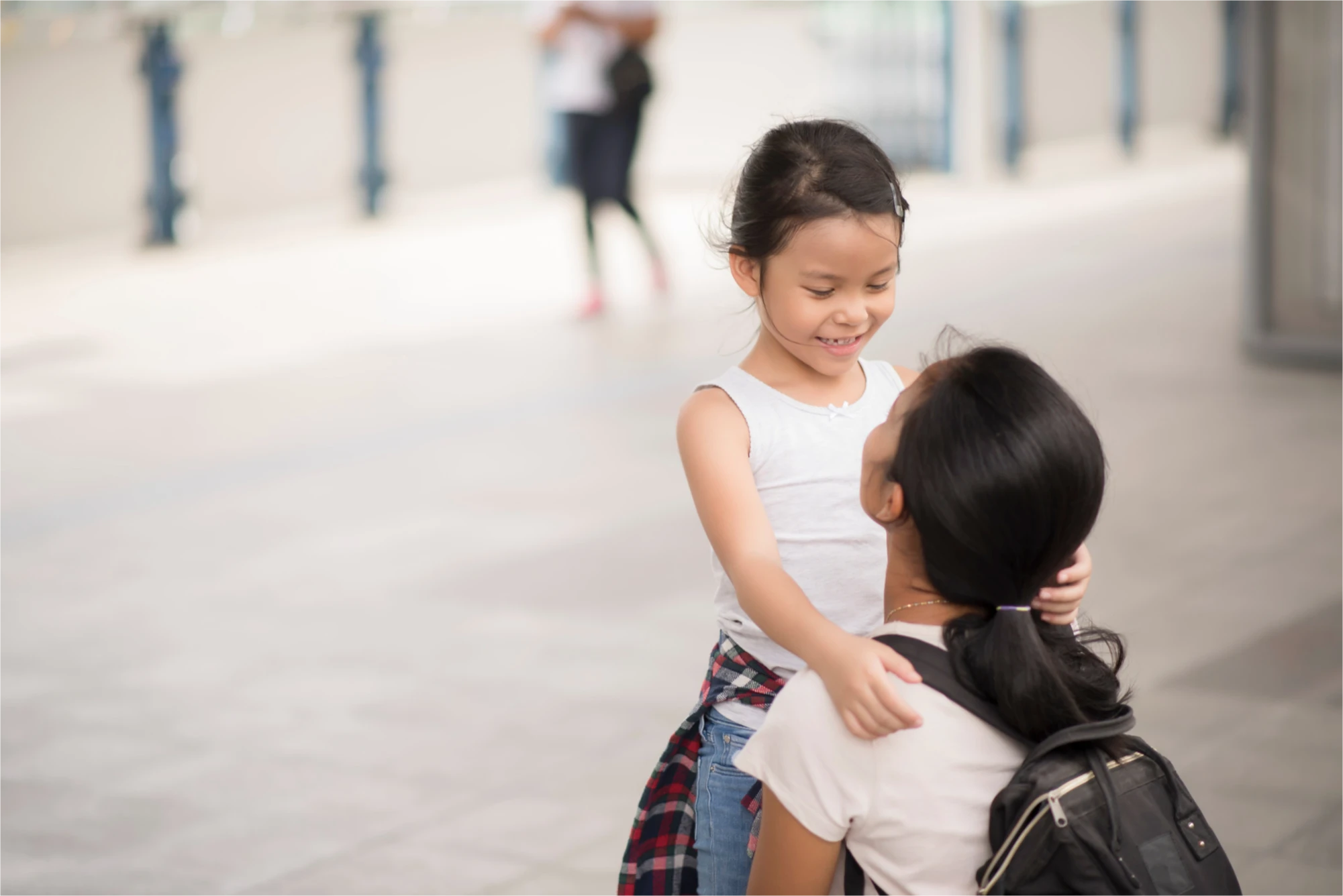 Young child in white top smiling at her caregiver during what appears to be a school drop-off moment.