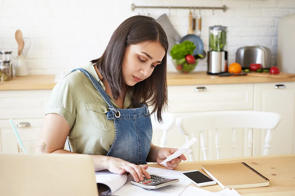 Woman in kitchen reviewing documents and using calculator at table