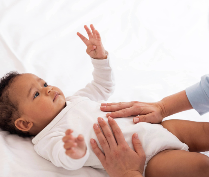 Bebé recostado en la cama mientras un adulto le toca el abdomen, acompañado de texto sobre consejos pediátricos para aliviar los gases en bebés.