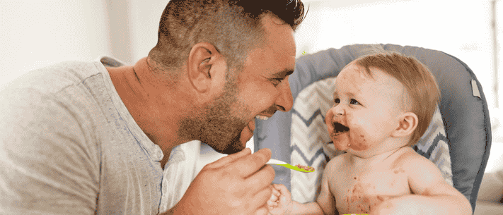 Father Feeding His Child in High Chair with Food-Covered Face and Torso