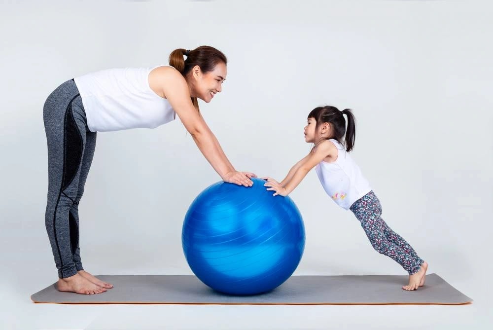 A Woman And A Young Girl Exercising Together, Leaning On A Large Blue Exercise Ball On A Gray Exercise Mat