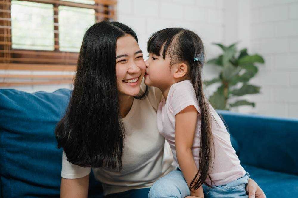 Child Leaning to Kiss Smiling Woman on Blue Couch Indoors
