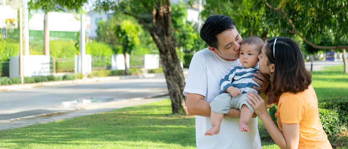 Family With Man And Woman Kissing Baby On Cheeks; Set Against Green Lawn And Trees
