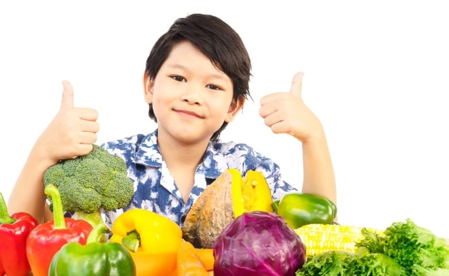 Smiling child giving thumbs up with various colorful vegetables in front. 