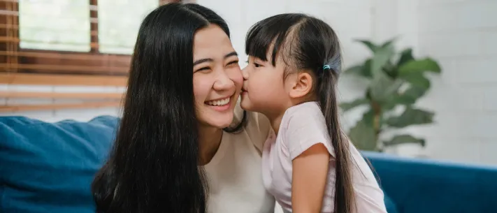 Young daughter kissing mother's cheek on couch in loving moment together