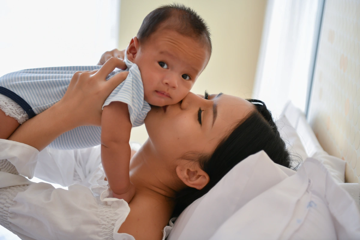 Mother kissing baby as she carries them while lying on bed.