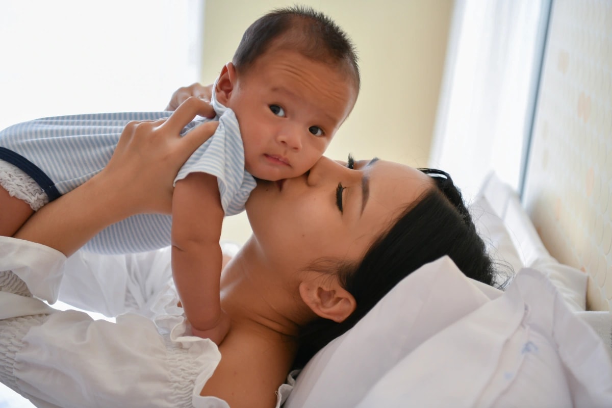 Mother kissing baby as she carries them while lying on bed.