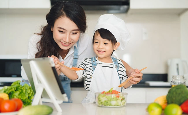 Mother And Child Smiling While Looking At A Tablet On Kitchen Counter, With Salad And Ingredients Nearby