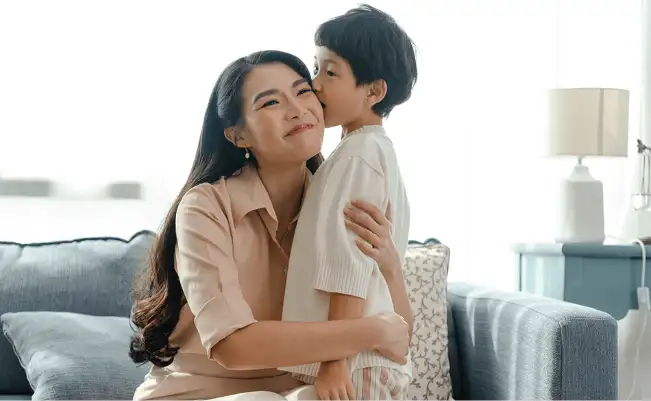 Mother sitting on sofa hugging son standing and kissing her on the cheek. 