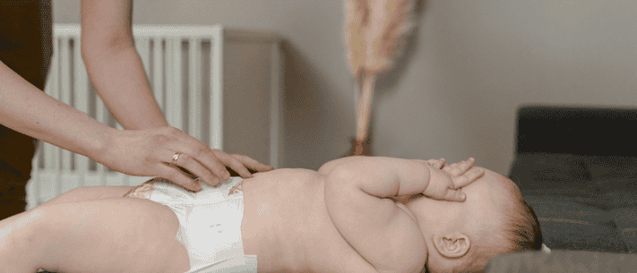 Baby Lying on Back Wearing Diaper with Adult's Hands Gently on Torso, in a Room with White Crib and Dried Floral Arrangement