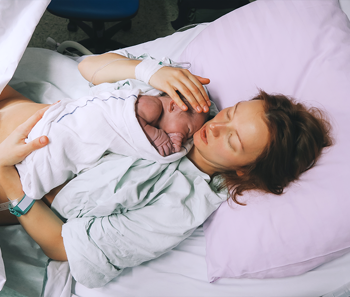 Madre descansando en la cama del hospital con su recién nacido en brazos tras el parto, reflejando recuperación y vínculo afectivo.