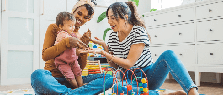 Two Women and Baby Playing on Floor with Colorful Toy in Nursery