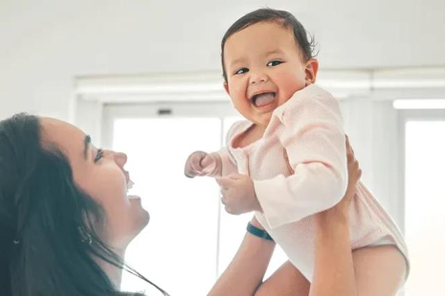 Mother Holding Up Smiling Baby in Light Pink Shirt, with Blurry White Background and Window