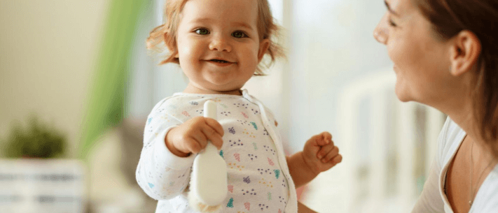 Smiling Baby in Patterned Onesie Holding Brush with Mother Nearby