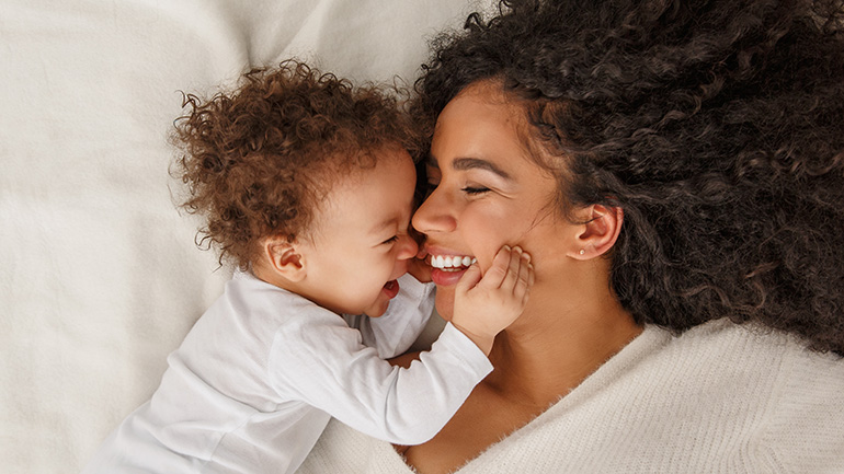 A smiling mother with curly hair lies down as her laughing baby affectionately touches her face.