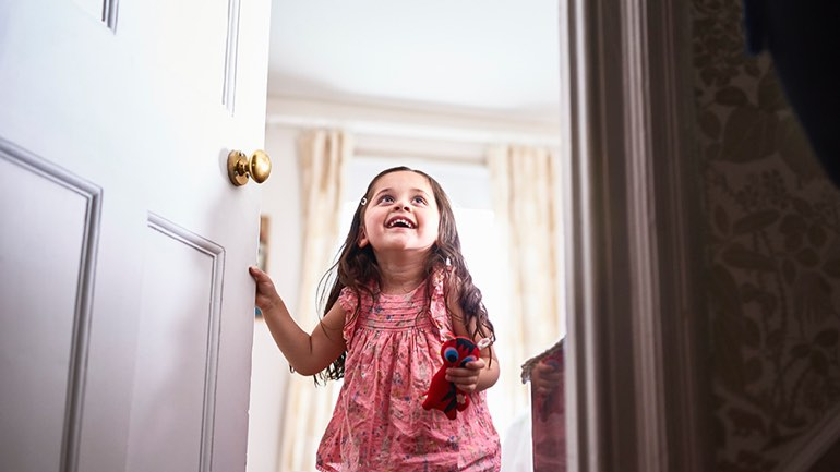 A smiling young child stands in a doorway holding a toy and looking upward with excitement.