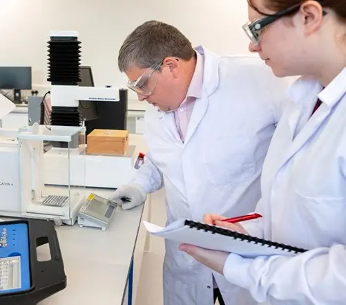 Two lab technicians measuring samples and recording data at a workstation.
