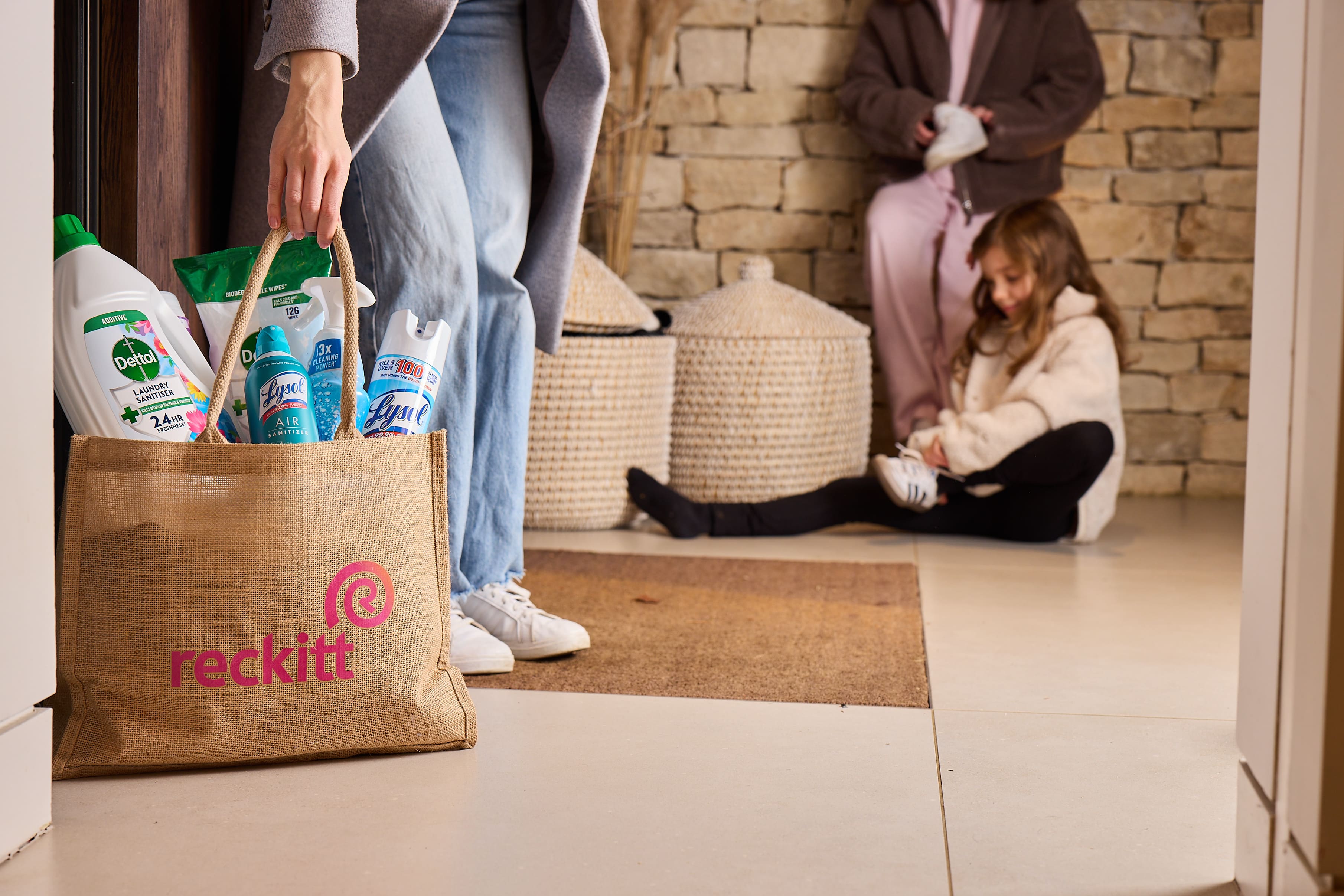 Person carrying a Reckitt-branded tote bag filled with household care products while family members prepare to leave home in the background