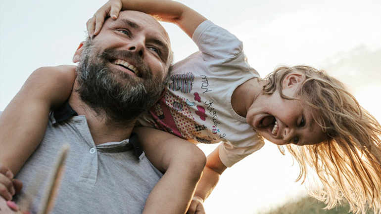 A laughing young girl enjoys a piggyback ride on the shoulders of her smiling, bearded father outdoors.