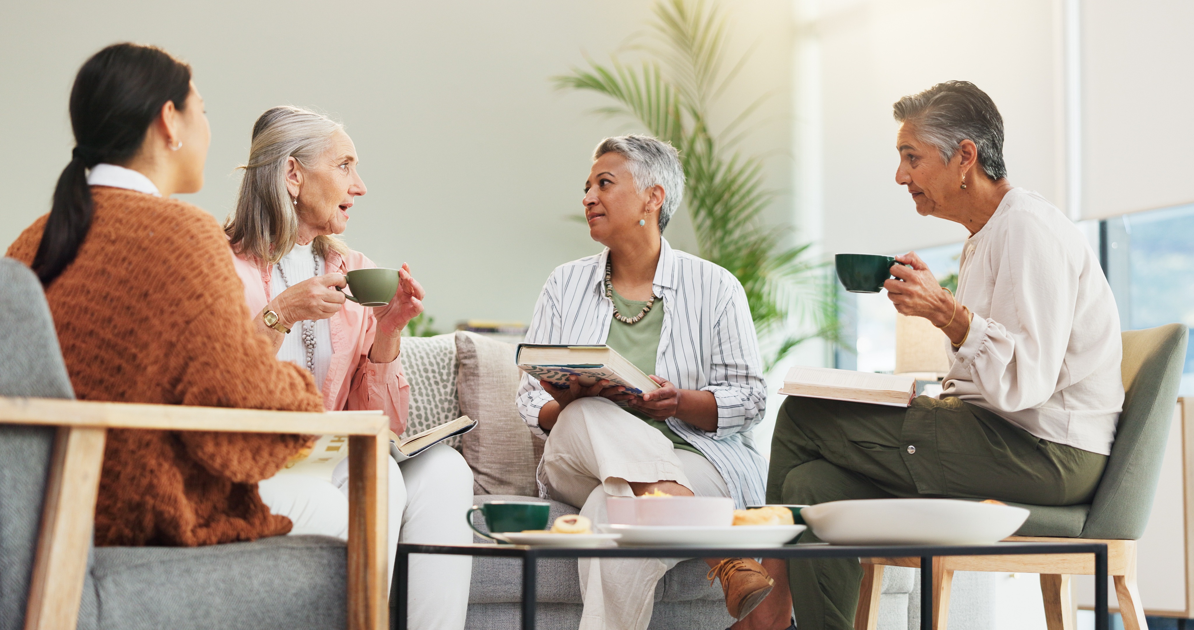 Four older women chatting over tea and books in a cozy living room.