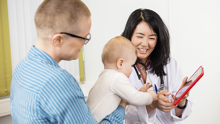 A smiling female doctor shows a digital tablet to a parent holding a baby during a medical appointment.