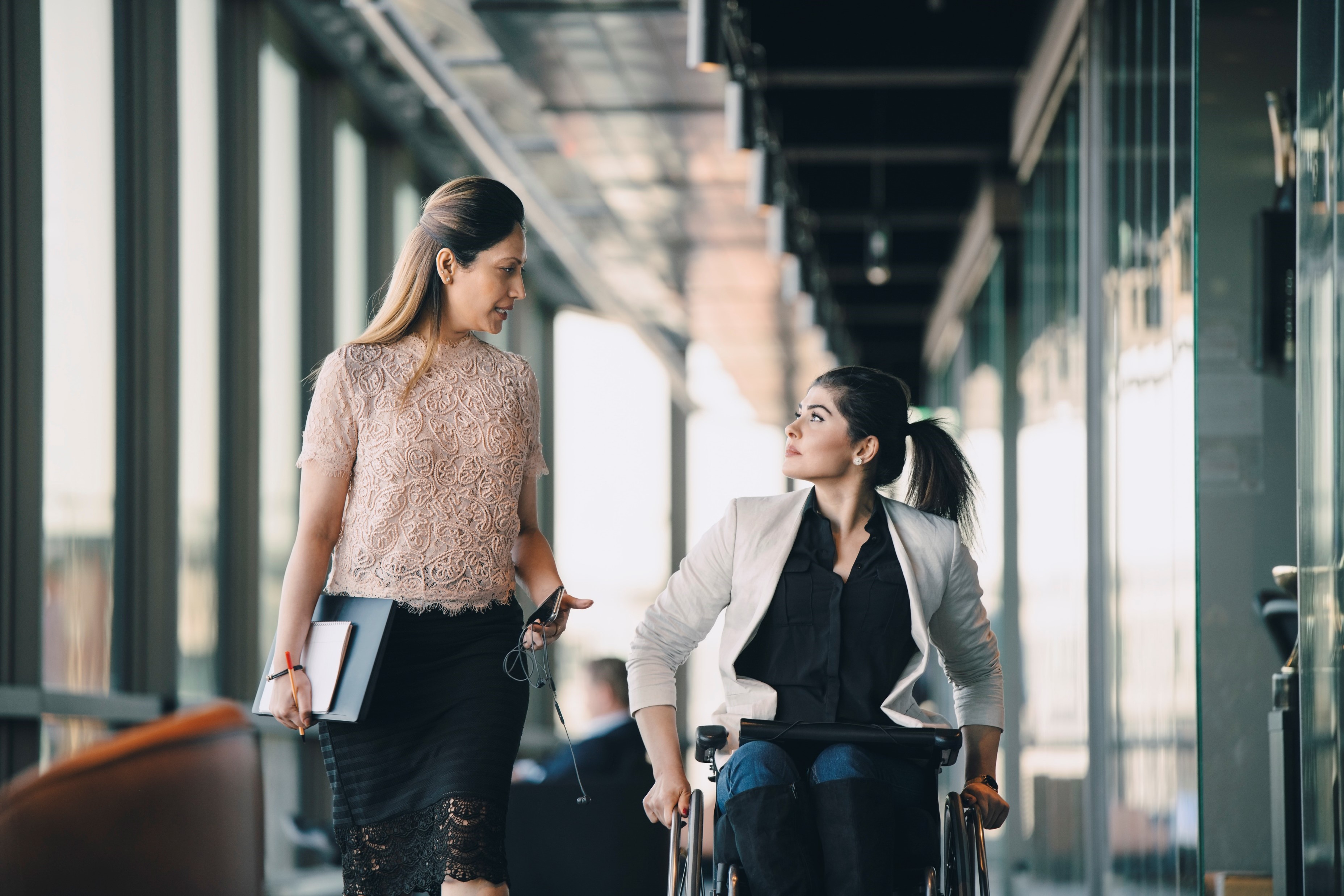 Two professionals walking and talking in an office hallway, including a person using a wheelchair