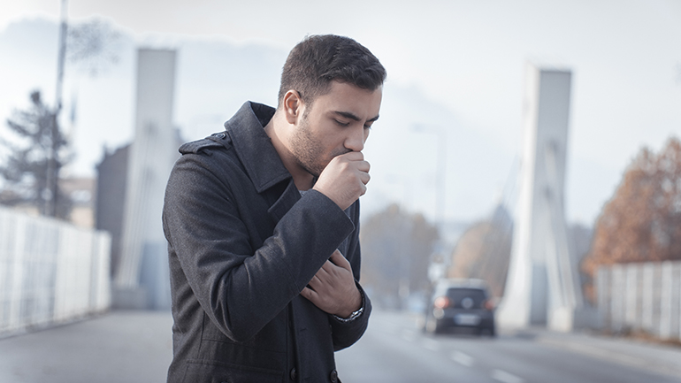 A man wearing a black coat coughs into his fist while standing outdoors near a bridge.