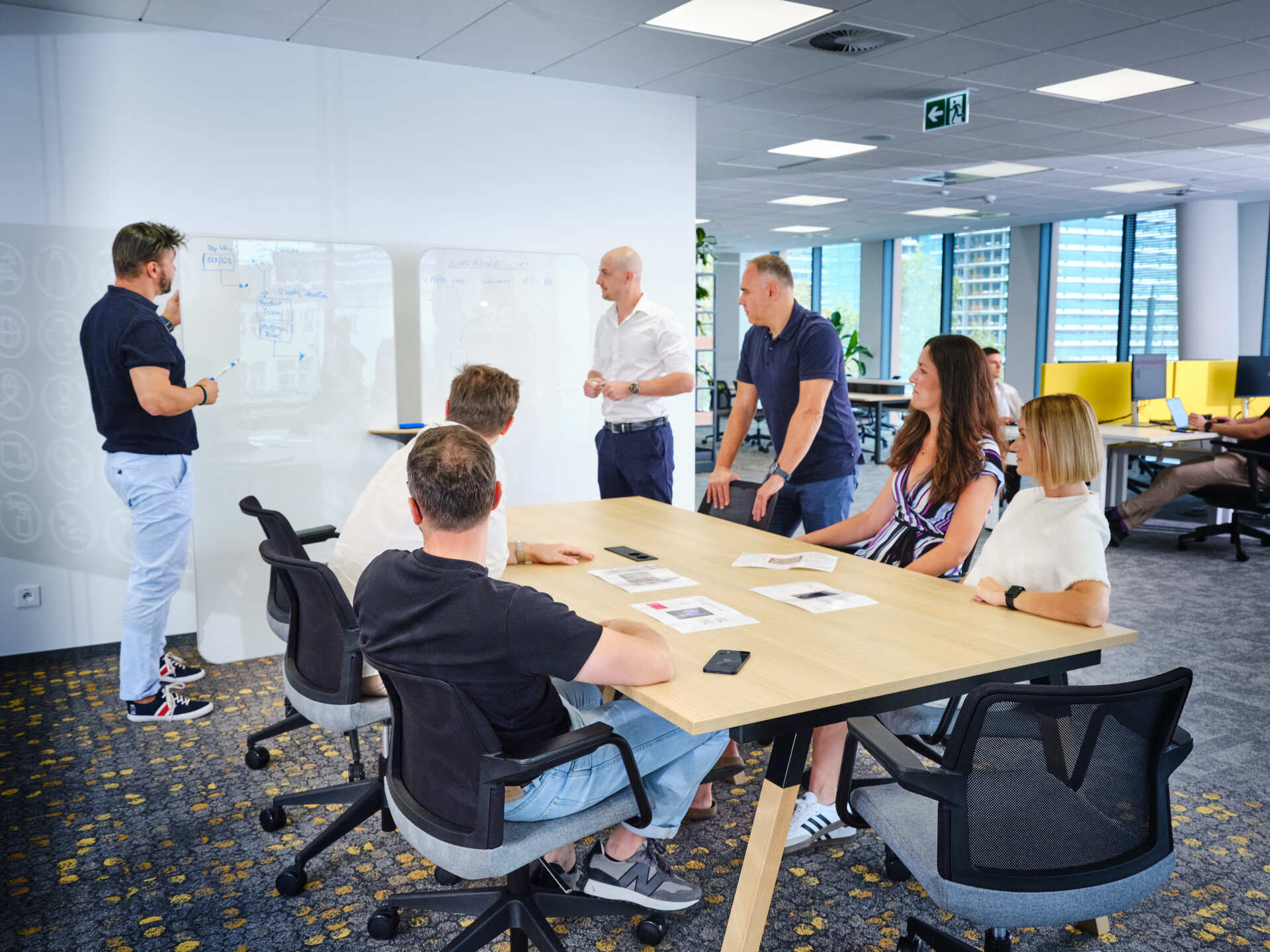 Team members collaborating in a meeting room, gathered around a table while presenting ideas on a whiteboard in a modern office setting