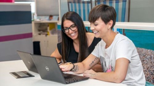 Two smiling female colleagues work together looking at a laptop screen in a modern office.