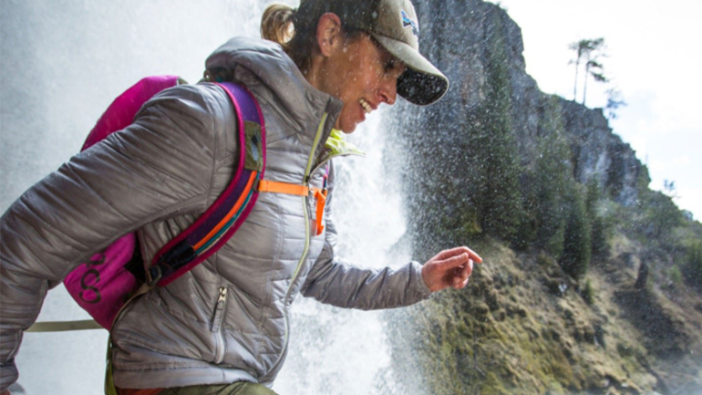 A smiling female hiker wearing a grey puffer jacket and baseball cap walks past a spraying waterfall in the mountains.