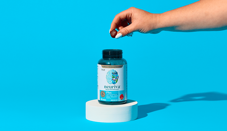 A person's hand holding a dark red gummy over an open bottle of Neuriva Plus Brain Performance supplements on a small white pedestal against a solid blue background.
