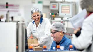 Two factory workers in hairnets and safety glasses smiling while packaging products.