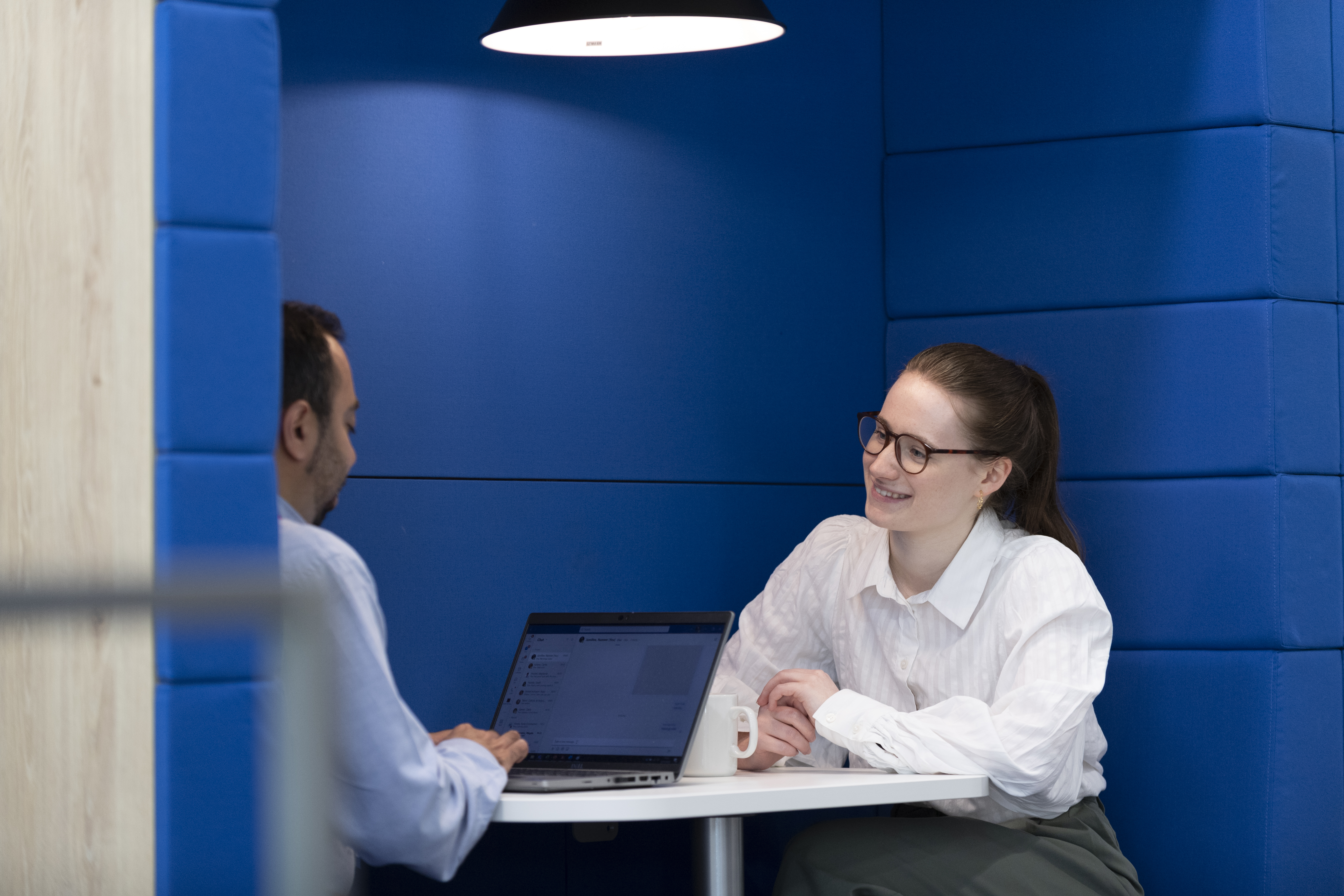 Two colleagues meeting in a private office booth, discussing work across a table with a laptop and coffee mug