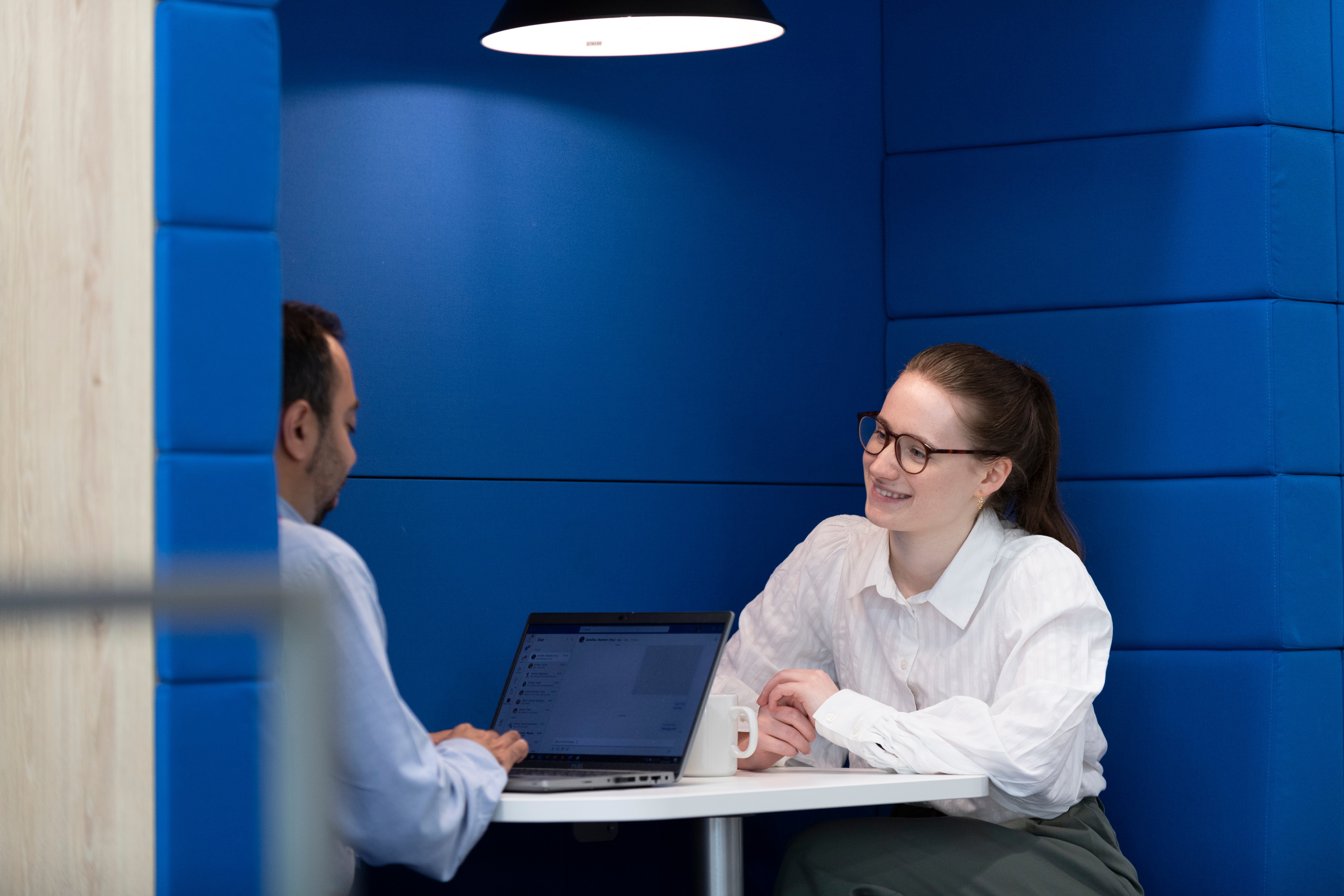 Two colleagues meeting in a private office booth, discussing work across a table with a laptop and coffee mug