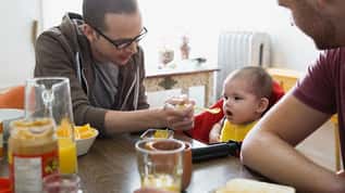Adult feeding a baby while seated together at a dining table with family members nearby