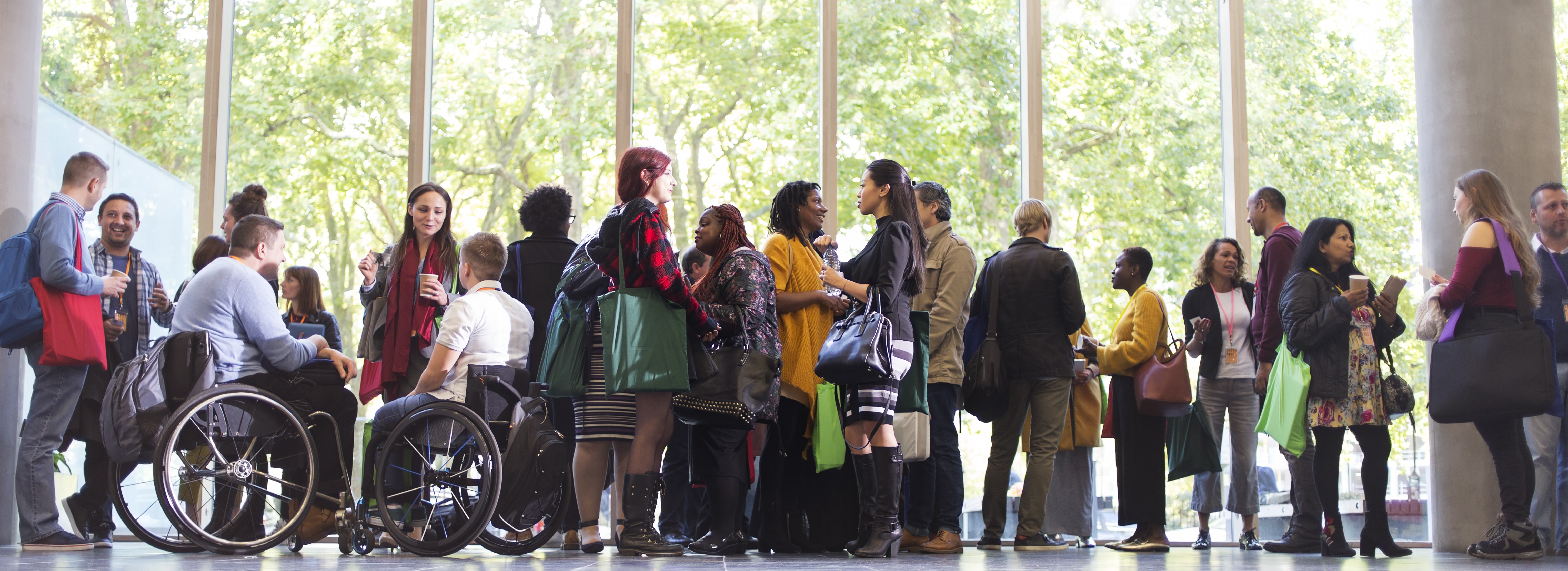 A diverse group of people socialize and network in a brightly lit office lobby with large windows overlooking green trees.