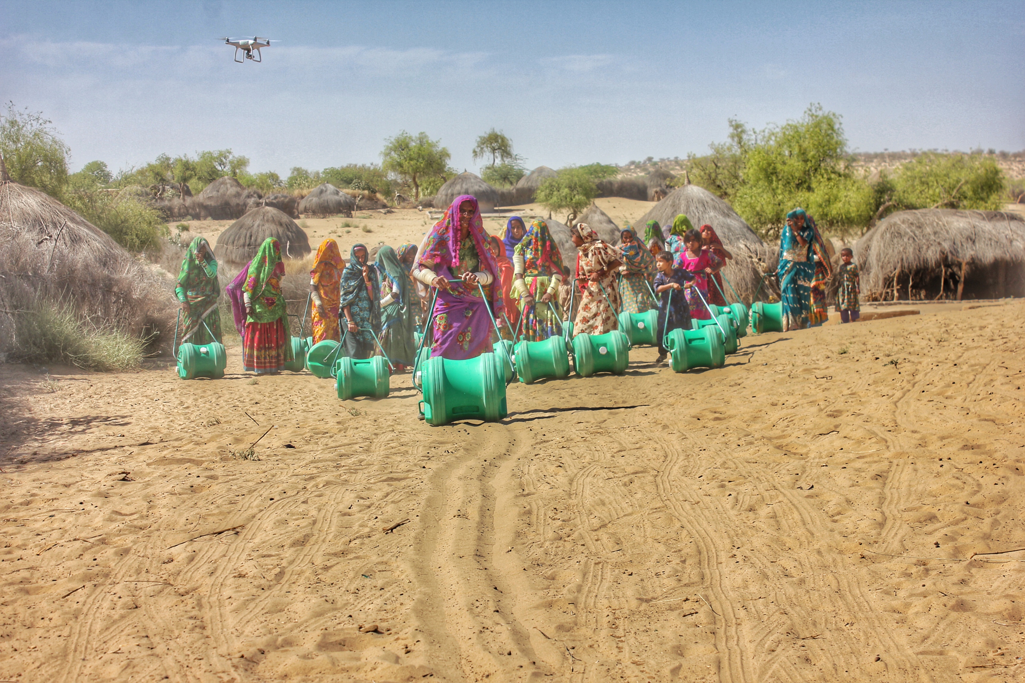 Group of women carrying water containers in a rural village with a drone overhead