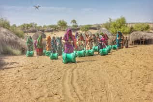 Group of women carrying water containers in a rural village with a drone overhead