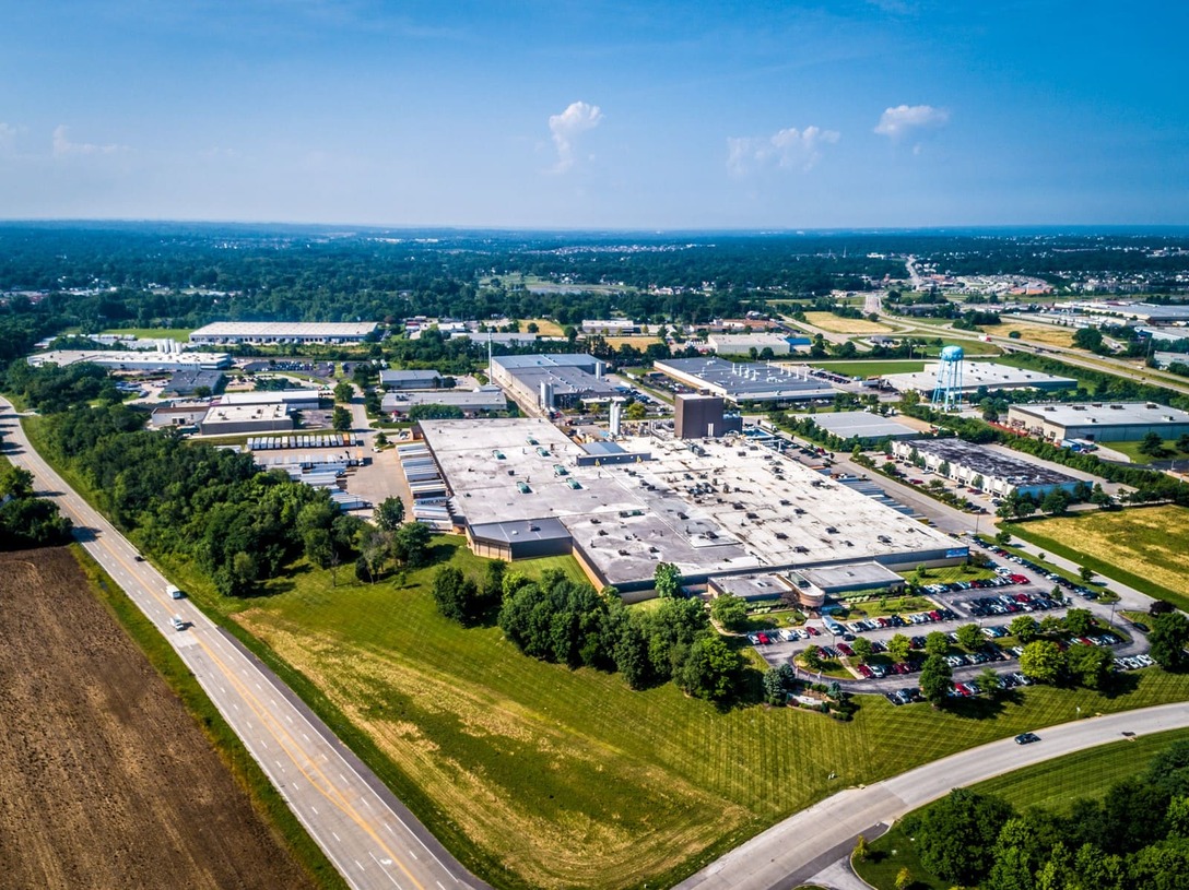 Aerial view of a large industrial complex surrounded by parking lots, roads, and greenery under a clear sky