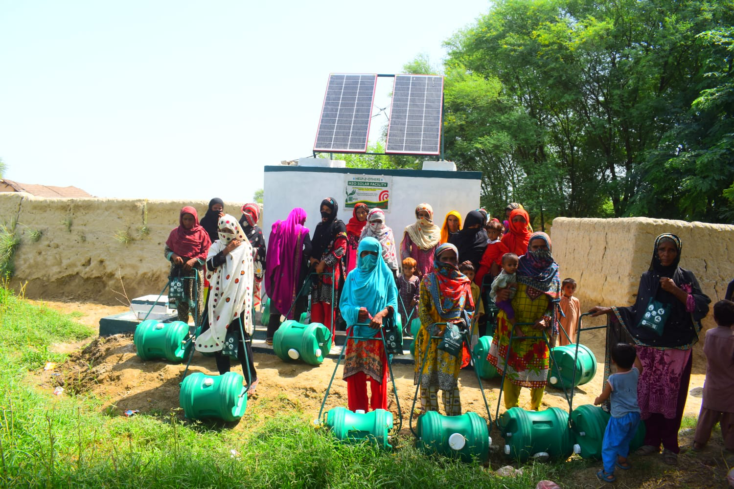 A group of women and children stand outdoors near a solar-powered water facility, each holding green water containers on wheels